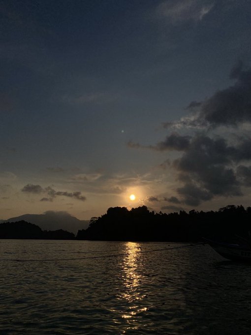 A serene sunset over water with silhouetted islands and a boat, the sun's reflection shimmering on the rippling sea.