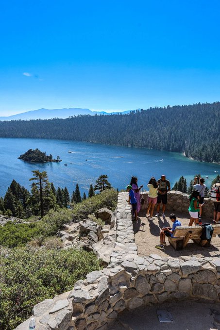 Tourists at a scenic overlook with stone walls taking photos of a stunning blue lake (Emerald Bay) surrounded by forested mountains under a clear blue sky, likely at Lake Tahoe.