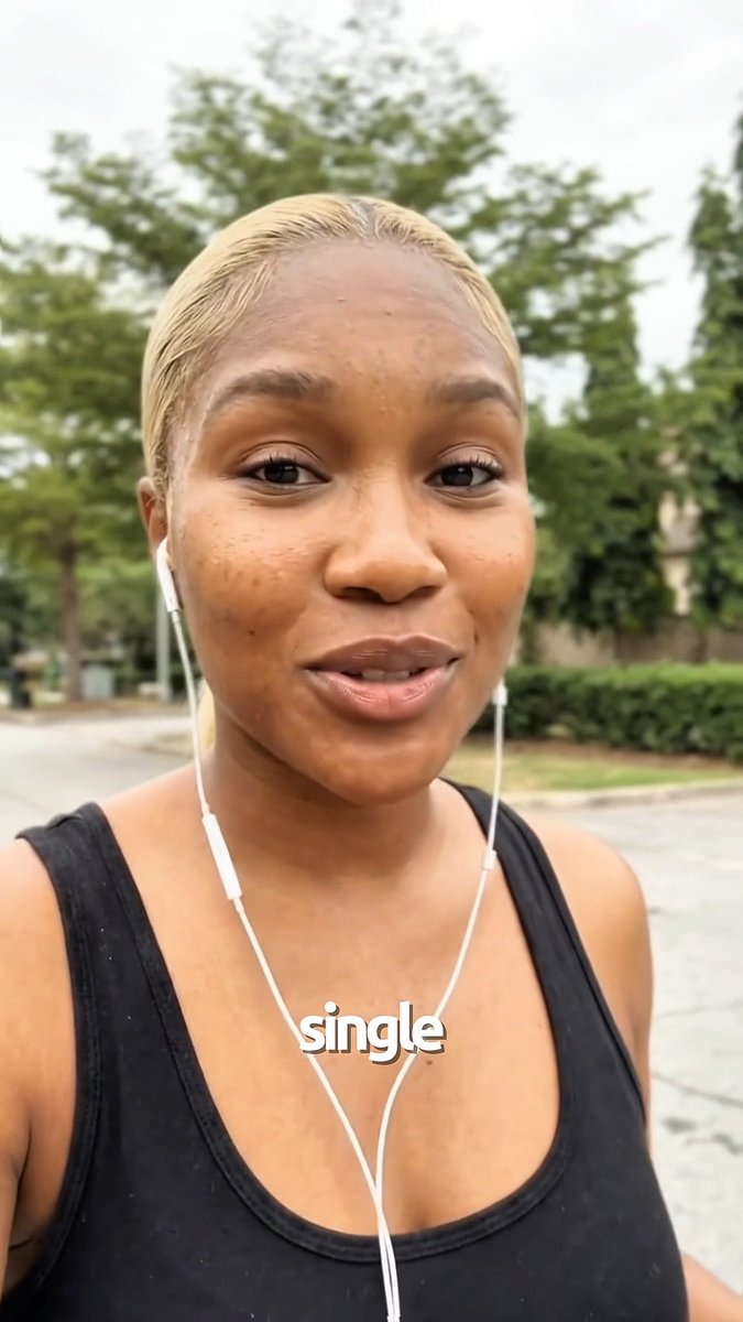 A young woman with short blonde hair wearing a black tank top and white earbuds takes a selfie outdoors, with the word 'single' displayed as text on the image.