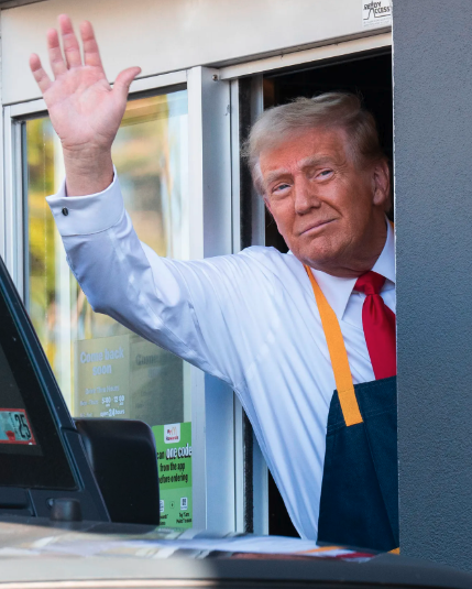 Former US President Donald Trump waves while wearing a blue shirt, red tie, and an apron at a drive-thru window.