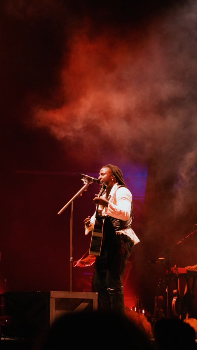 A musician with dreadlocks, wearing a white shirt and dark vest, performs on stage while playing an acoustic guitar and singing into a microphone amidst red lighting and smoke.