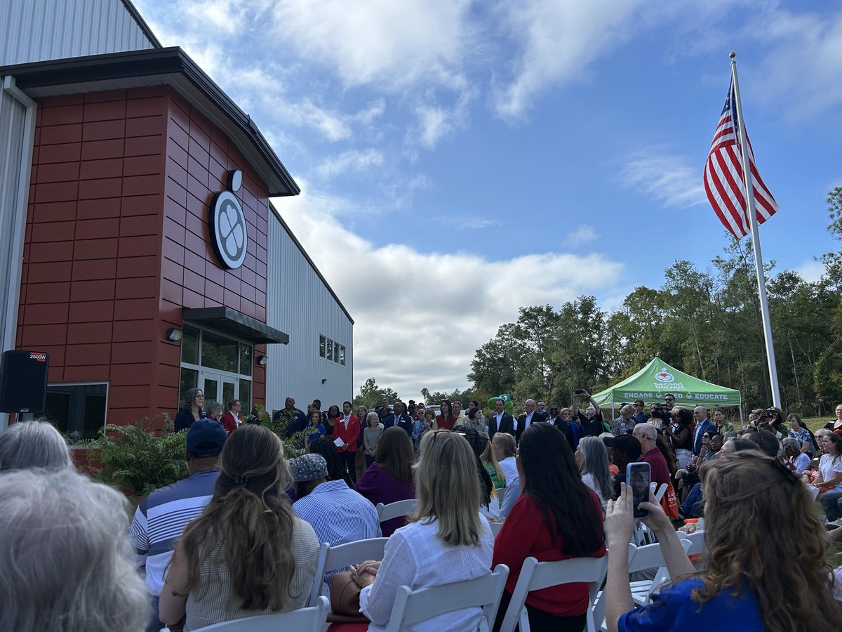 An outdoor ceremony is taking place beside a modern building, with a seated crowd facing a group of people near the entrance. An American flag flies above them, and a green event tent is visible nearby. There is text on the building and tent.