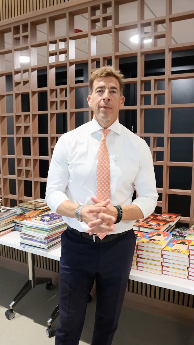 A man in a white shirt and orange patterned tie stands with hands clasped in front of wooden shelving, with stacks of colorful children's books on tables beside him.
