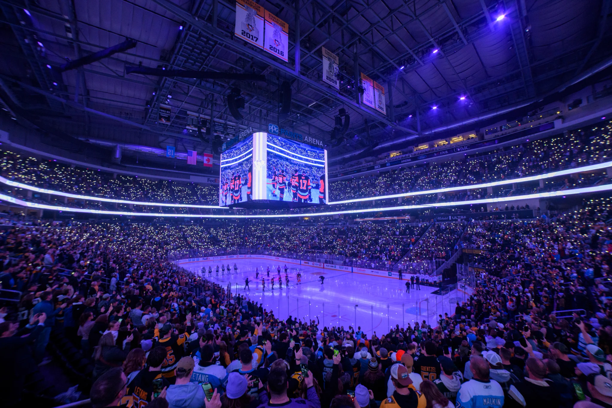 A large indoor ice hockey arena, filled with spectators. The crowd is illuminated by small lights, creating a vibrant atmosphere. The ice surface is brightly lit, with players lined up and ready for the game. Overhead, a large digital screen displays an image related to the ongoing event. The arena is adorned with banners and flags.