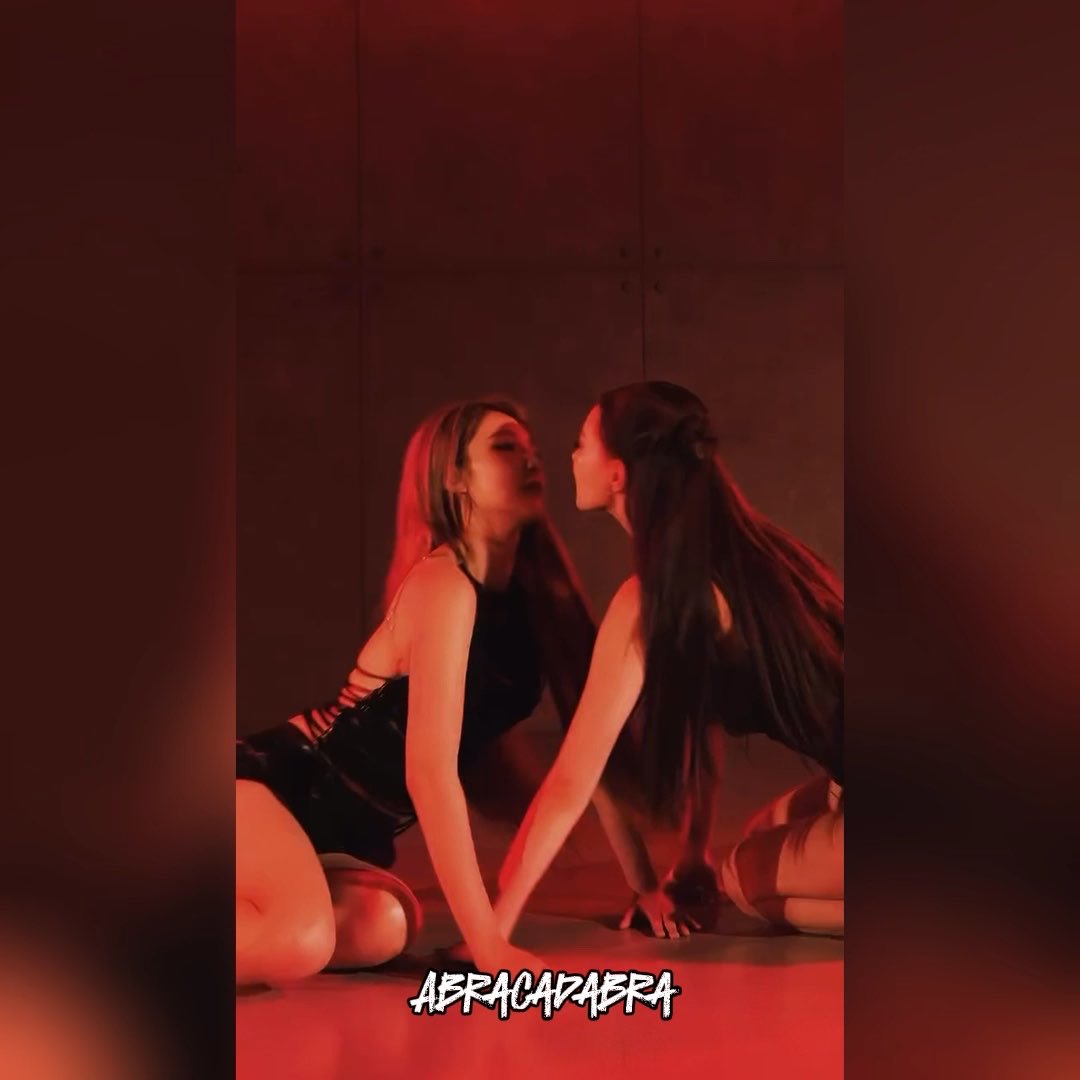 Two women in dark outfits pose intimately in a dimly lit room with red lighting, with the text 'ABRACADABRA' displayed at the bottom.