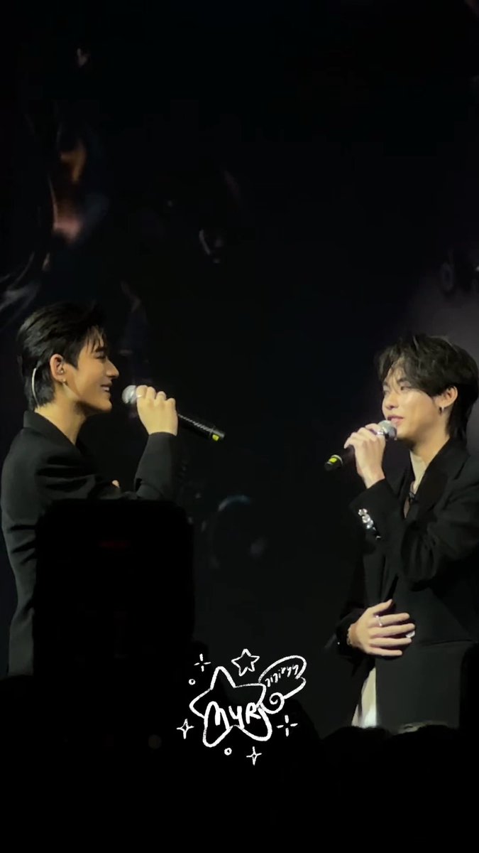 Two male performers in dark suits hold microphones on a dimly lit stage during a concert, with a white star logo and the Korean text '아이돌' (Idol) visible at the bottom.