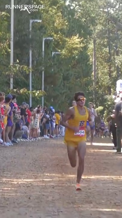 A male runner wearing a yellow tank top and sunglasses competes on a dirt path during a cross-country race, with spectators lining the sidelines under tall trees, and a 'RUNNERSPACE.com' watermark in the corner.