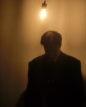 Moody, low-lit photograph of a silhouetted person in a dark suit standing against a wall illuminated by a single bare lightbulb.