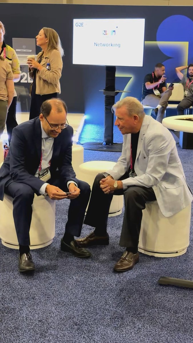 Two men sit on white stools having a conversation at what appears to be the G2E Networking event, with other attendees visible in the background.