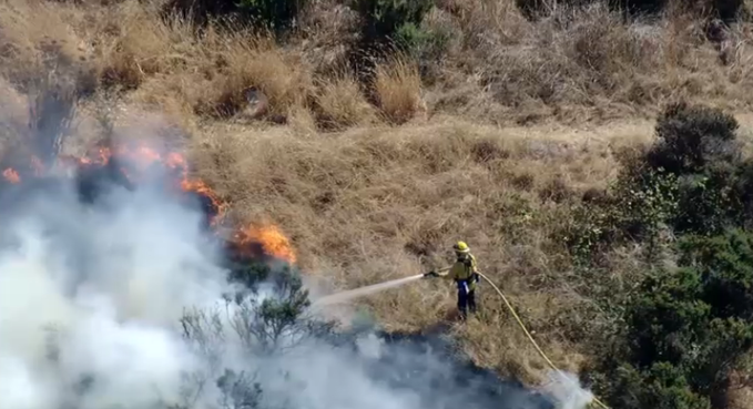 A firefighter in protective gear spraying water from a hose onto a brush fire burning on a dry, grassy hillside.