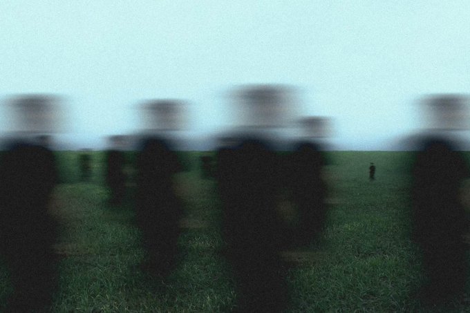 Blurry, atmospheric image showing dark silhouettes of figures or objects standing in a grassy field under a pale, overcast sky.