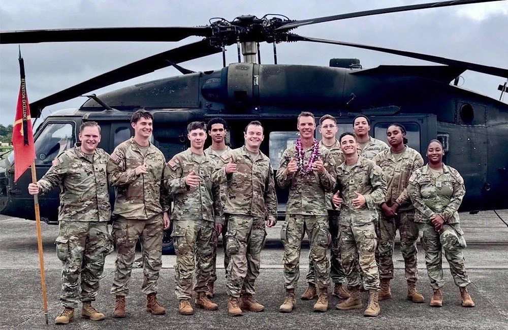 A group of 11 people in U.S. Army uniforms pose together in front of a military helicopter. One person holds a guidon while another wears a purple flower lei.