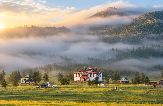 A serene landscape at sunrise showing a traditional Buddhist monastery with golden roofs nestled in a misty valley, surrounded by forested mountains and a grassy meadow.