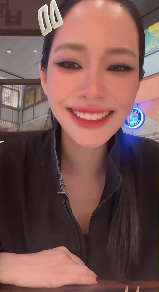 A smiling woman with dark hair pulled back wears a black zip-up jacket in what appears to be a restaurant setting, with a small white object visible at the top of the frame.