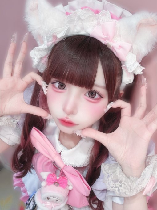 A person in elaborate pink and white frilly cosplay attire with cat ear headwear, dramatic makeup, long decorated nails, and a small plush accessory on their outfit, posing with hands framing their face.