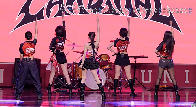 A K-pop girl group performing on stage with their backs to the camera, all raising one arm in unison, wearing black and red outfits; an October 2025 calendar is overlaid at the top.