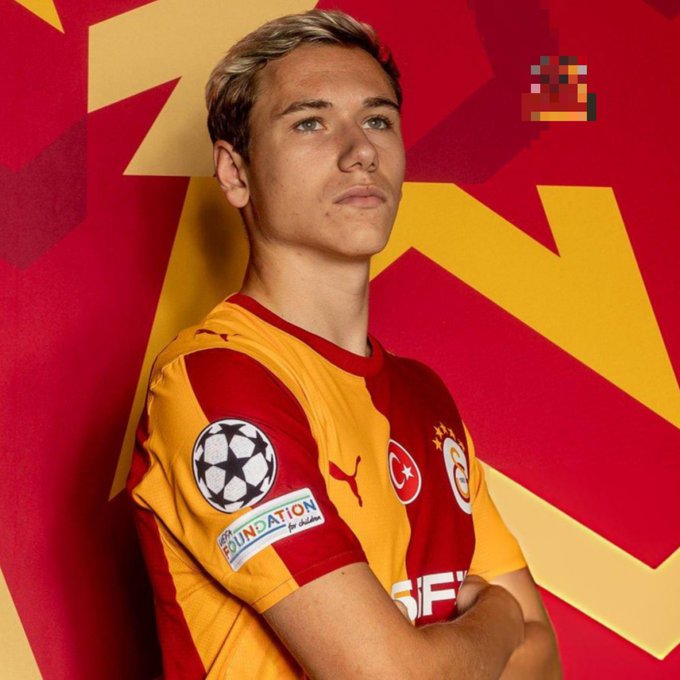 A professional portrait of a young soccer player with blonde hair wearing a red and yellow Galatasaray jersey featuring the Champions League patch, posed against a red and yellow background.