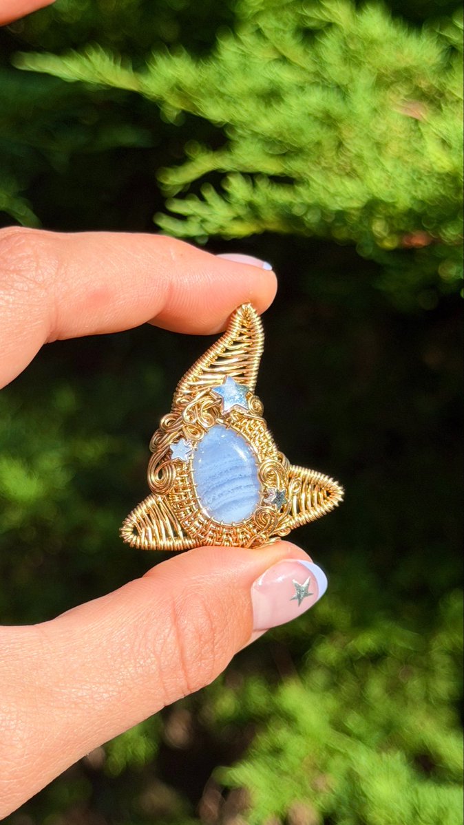 A person's hand, with star-painted nails, holding a handcrafted pendant made of wire-wrapped light blue stones and small star accents, with evergreen foliage blurred in the background.