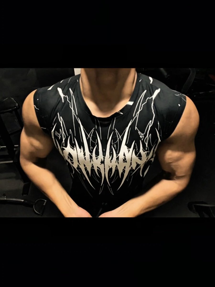 A muscular man wearing a black sleeveless shirt with white graphic text sits in what appears to be a gym.