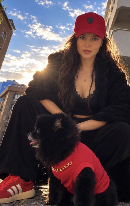 A woman wearing a red Raiders cap, black coat, and red Adidas shoes, posing at sunset next to a black poodle dressed in a matching red outfit with a gold chain.