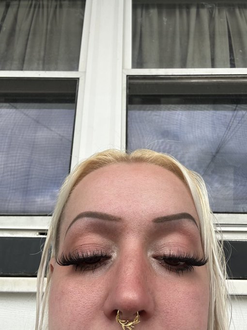 A close-up selfie of a blonde woman showing her eyes with long false eyelashes and a gold septum nose ring, with windows visible in the background.