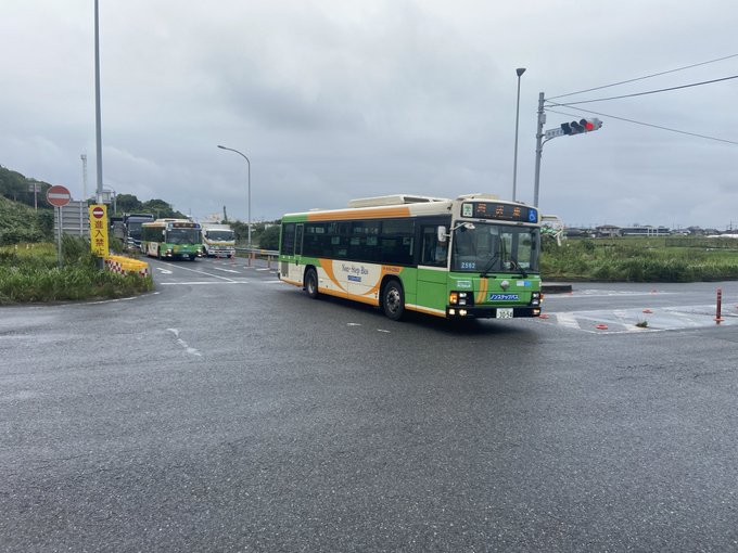 A green and orange Toei Bus marked "Non-Step Bus" navigates a roundabout on an overcast day, with the surrounding tropical scenery and the caption indicating it has just crossed the Aqua-Line into Chiba Prefecture via the Uchibo Line route.