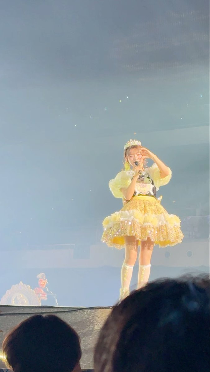 A performer on stage in a yellow frilly idol outfit with a tiara, holding a microphone and saluting the audience, with a small mascot visible to the left.
