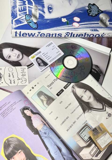 K-pop merchandise spread out, featuring albums and photobooks related to NewJeans including 'Hanni' and 'Haerin' visible, along with a CD, stickers, and an ID-style photo card.
