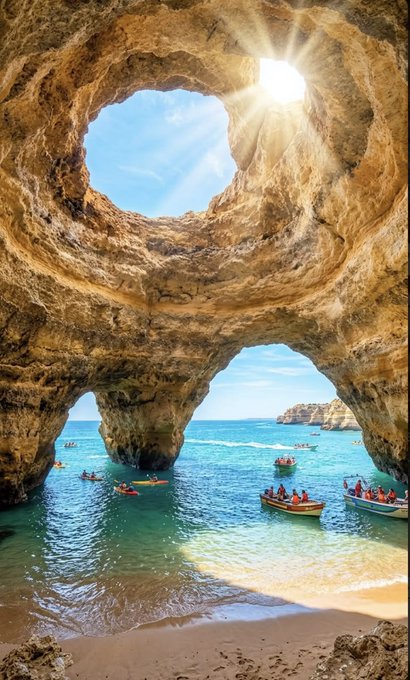 A scenic view from inside a large sea cave featuring a circular opening in the ceiling that lets sunlight stream down onto turquoise waters filled with small boats, kayaks, and swimmers near a sandy beach.