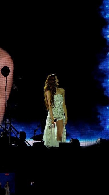 A female performer in a white dress standing on a dimly lit stage holding a microphone with blue lights in the background.