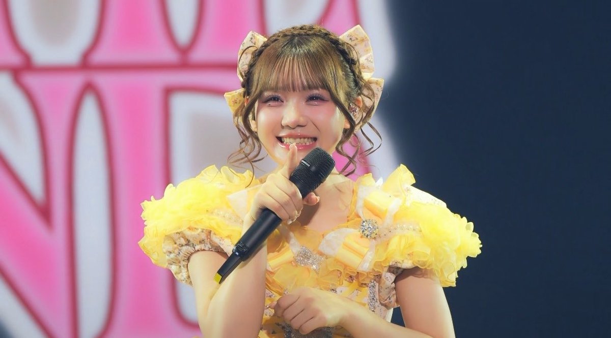 A smiling young woman with curly brown hair and decorative bows, wearing a ruffled yellow outfit and holding a microphone close to her mouth during a stage performance.