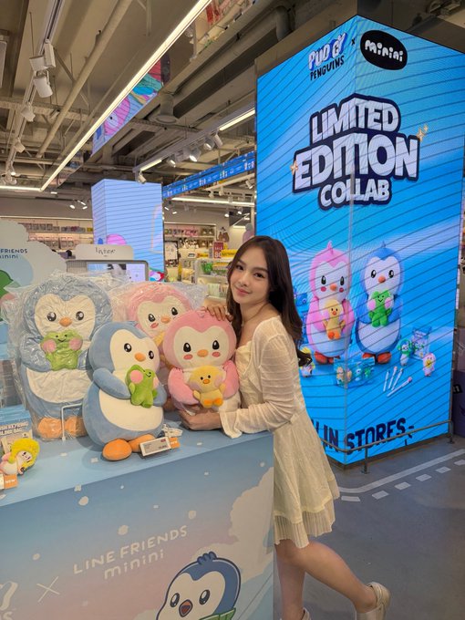 A young woman in a white dress poses at a "LINE FRIENDS mini" and "minini" limited edition collaboration display in a store, with several penguin plush toys arranged on a blue stand.