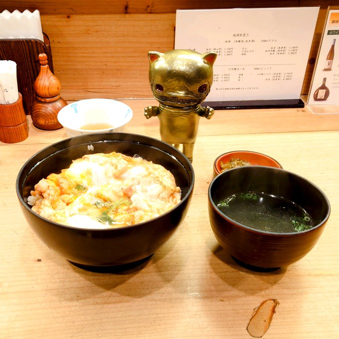 A Japanese meal featuring a bowl of rice topped with egg and seafood, accompanied by a bowl of miso soup, with a golden cat figurine standing behind them.