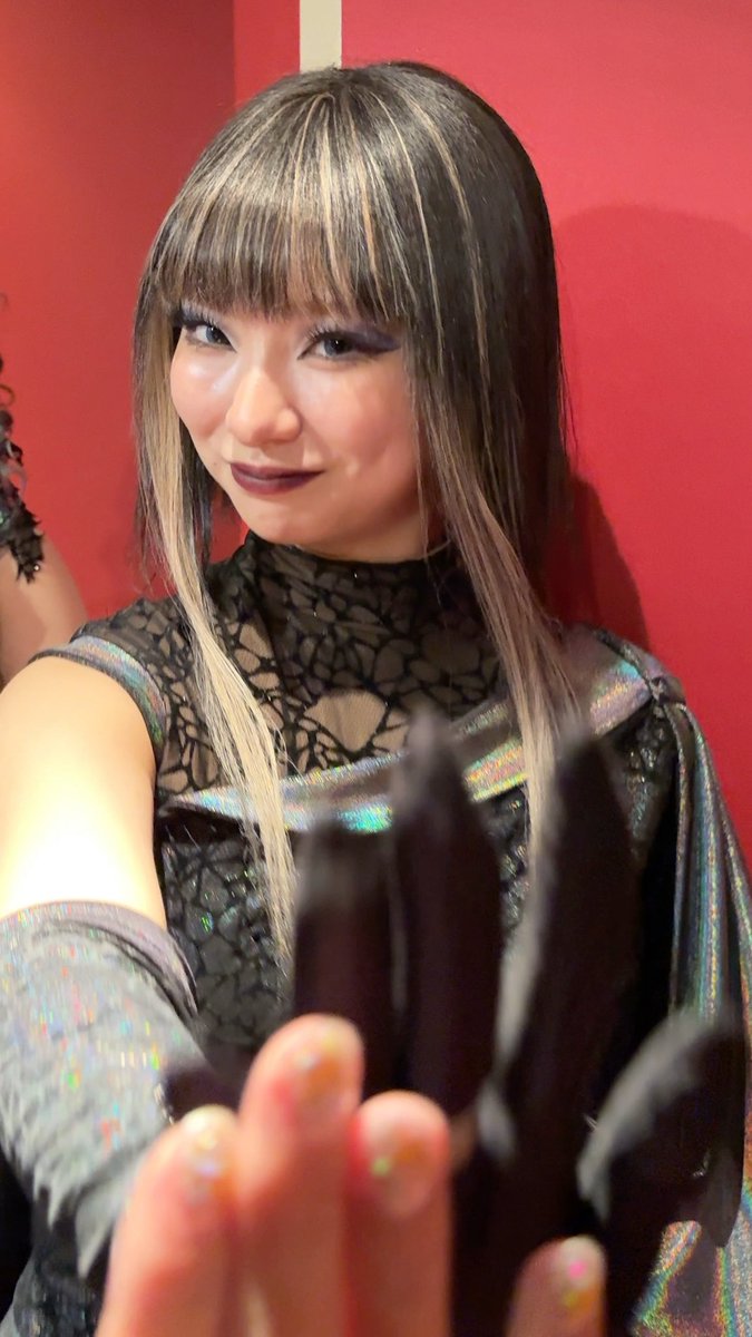 A woman with blonde-streaked bangs, heavy eye makeup, and a black lace outfit poses against a red background while holding up a black-gloved hand.