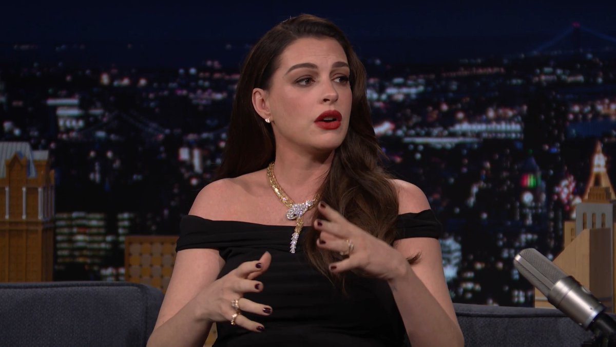 Anne Hathaway appears on The Tonight Show with Jimmy Fallon, wearing an elegant black off-shoulder dress and a statement gold necklace with diamond accents, gesturing expressively while seated across from a microphone. The image relates to the post as it captures Hathaway during her late-night interview appearance, which likely included discussion of her red carpet photography at the film's premiere.