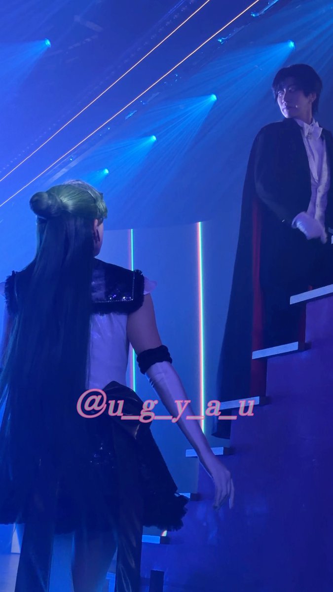 A stage scene with blue lighting shows a person in a dark cloak standing on elevated stairs, while another person with long hair in a costume looks up at them; the watermark '@u_g_y_a_u' is visible.