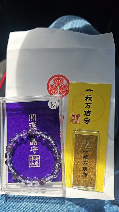 Two Japanese amulets, known as omamori, are displayed: a purple one with a crystal bracelet reading '開運 晶守' and a gold one reading '一粒万倍守'.