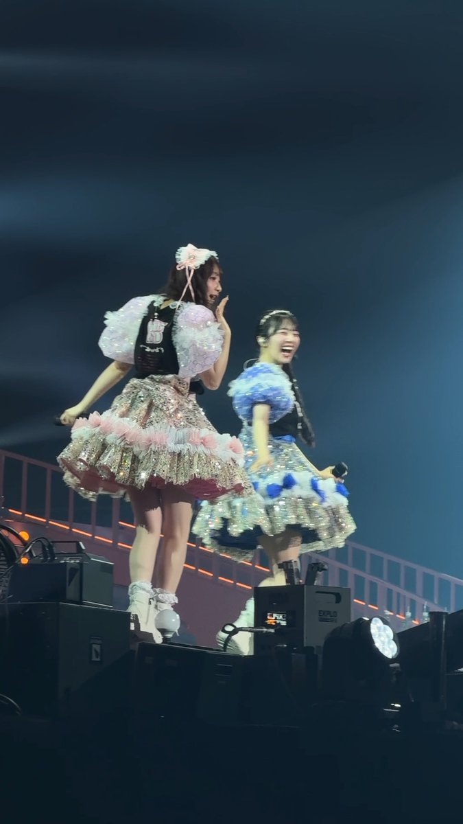 Two Japanese idols in elaborate frilly pink and blue stage costumes perform on stage at night, captured mid-performance with microphones.