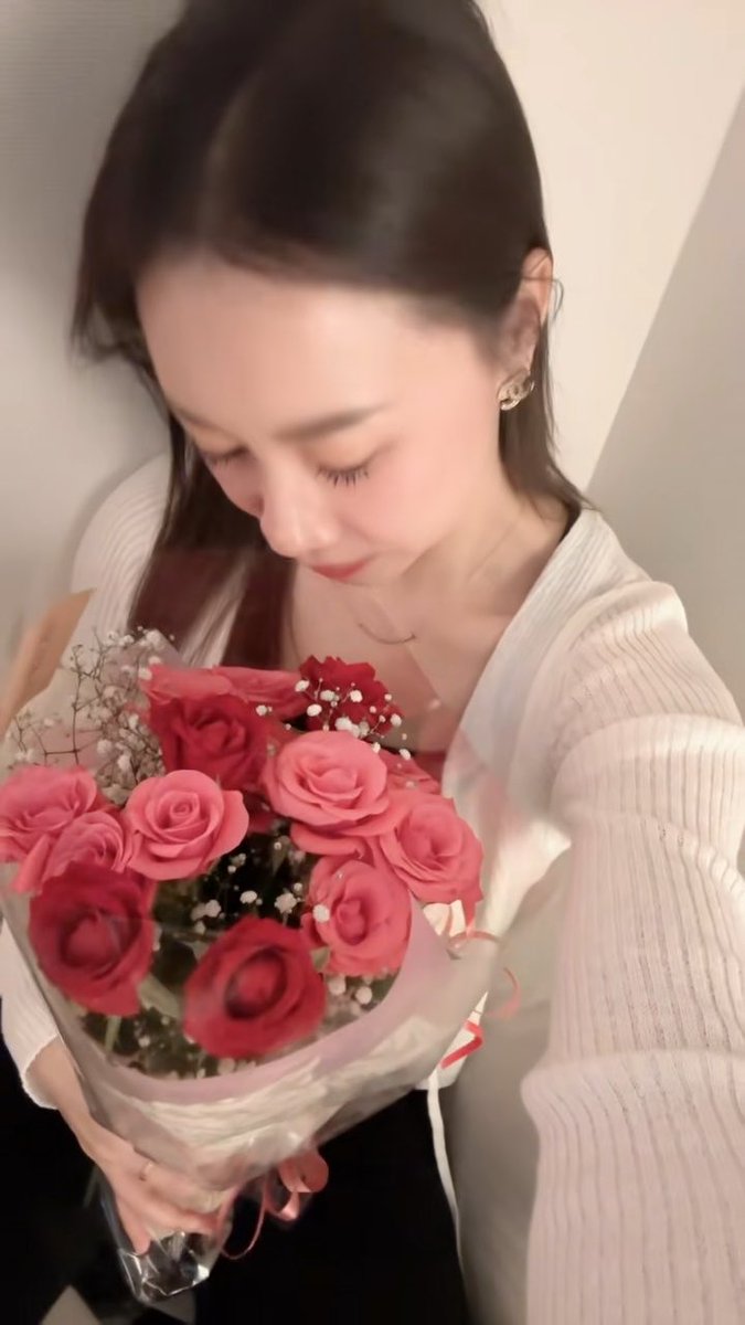 A young woman with long dark hair wearing a white cardigan looks down at a bouquet of red and pink roses she is holding.