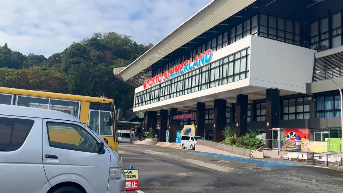 A photograph of the Tokyo Summerland entrance building with its red and blue signage, featuring a parking lot with a yellow bus, white van, and small silver car visible in the foreground.