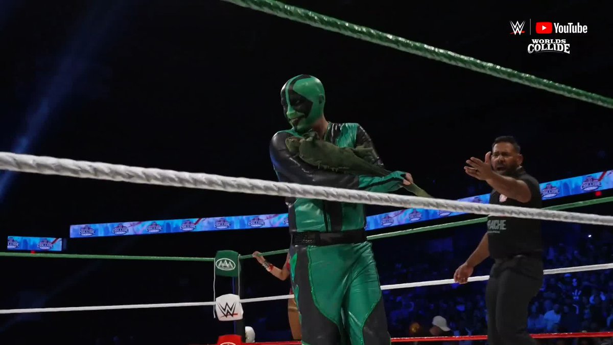 A masked wrestler in a green and black lizard-themed costume poses inside the ring, holding what appears to be a green prop, while another wrestler in black reaches toward him. The image features "WWE Worlds Collide" and "YouTube" branding in the top right corner, matching the caption announcing "La Yesca" (known as The Lizard) at the WWE Worlds Collide event.
