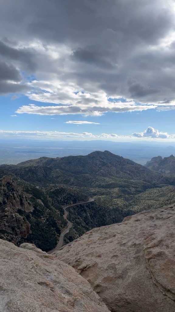 A scenic view from a rocky mountain overlook, showing a winding road cutting through forested hills and a distant city valley under a dramatic cloudy sky.