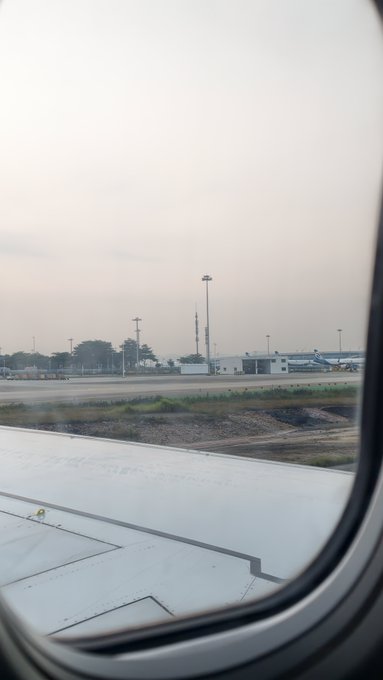 View from an airplane window showing the aircraft's wing and an airport tarmac with light poles and buildings at dusk.