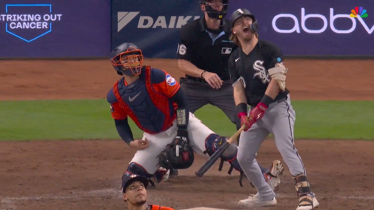 A Chicago White Sox batter with long hair celebrates with his head tilted back and mouth open after a hit, while the Houston Astros catcher in red and blue gear looks on, with the NBC logo and 'Striking Out Cancer' signage visible in the background.