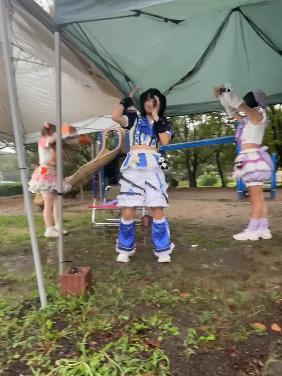 Several young women in colorful, frilly idol costumes are dancing and posing under a large green canopy tent at an outdoor location.