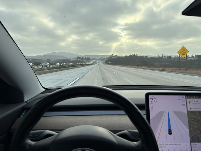A view from inside a Tesla driving on a highway with a 'TRAFFIC MERGE LEFT' sign visible; the dashboard display shows a speed of 76 mph and indicates the destination as 'Foothill Blvd'.