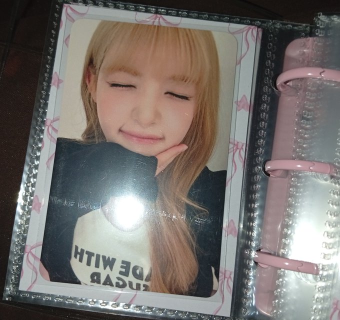 A photocard of a smiling blonde girl in a black and white 'WE WITH USADA' shirt is displayed in a clear plastic binder with a sparkly gem border.