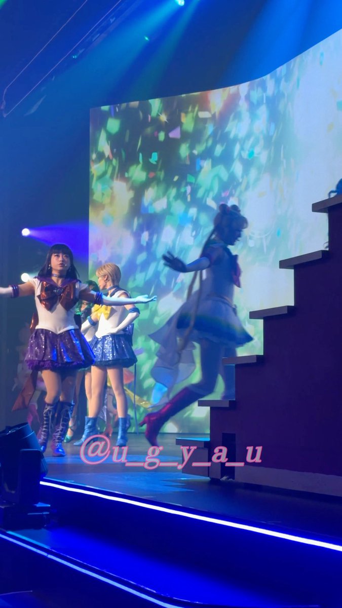 Three performers in sailor-style idol costumes perform on a stage illuminated by blue lighting and a colorful projected backdrop, with a watermark reading '@u_g_y_a_u' visible across the bottom.