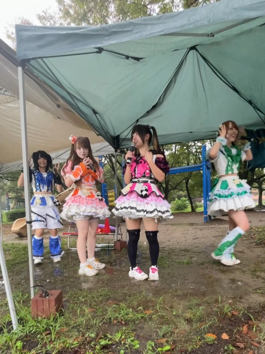 Four young women in colorful, frilly stage costumes perform under a green pop-up tent in an outdoor park setting, with microphones in hand.