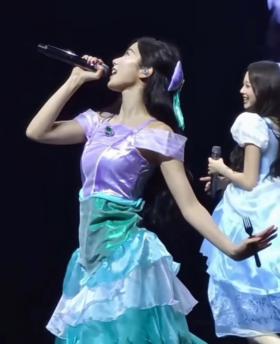 A young woman with long dark hair sings into a microphone on stage wearing a purple and teal princess-style gown, with another performer in a blue and white dress smiling in the background.